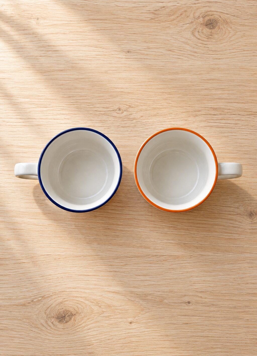 Twee witte koffiekopjes naast elkaar van bovenaf, de ene met een blauwe rand en de andere met een oranje rand.