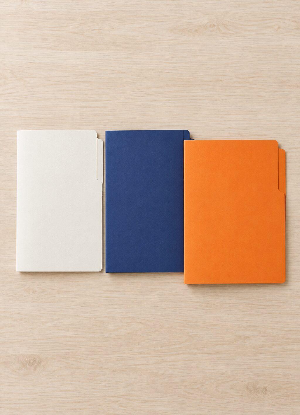 Trois chemises fermées en blanc cassé, bleu et orange côte à côte sur une table en bois, vues de haut.