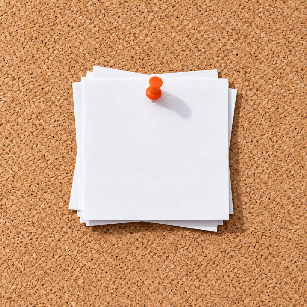 Stapel witte memoblaadjes met een oranje punaise op een kurkbord, van bovenaf.